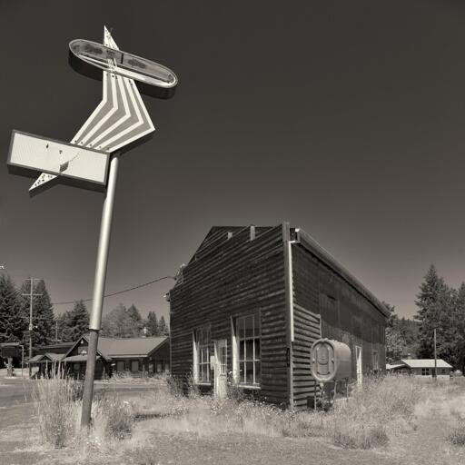 A small, vacant, clapboard store in a rural setting. It is surrounded by weeds. To the left is a disproportionately large Googie-style arrow-shaped sign on a pole.