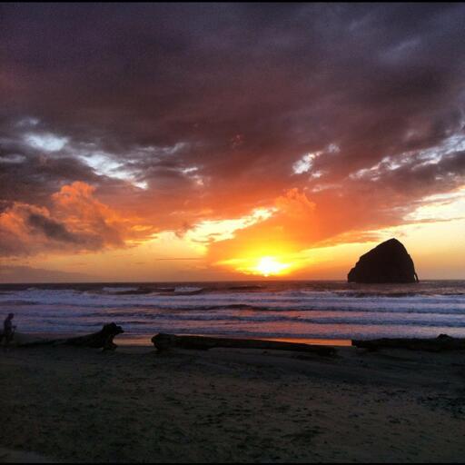 The sun sets through the clouds, seen from the beach with ocean waters lapping on shore. A huge rock is just offshore, and that rock has a natural little "handle" on its right side.