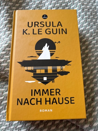 Ein Foto der Hardcover Ausgabe von Immer nach Hause von Ursula K. le Guin. Das Cover ist ockerfarben mit einer schwarzen und weissen Illustration in der Mitte, ein Wolf heult vor dem Mond und darunter ist ein auf dem Kopf stehendes Haus abgebildet, es sieht aus wie eine Spiegelung in einem See 